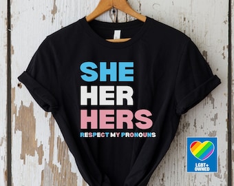 Könnte beinhalten: Schwarzes T-Shirt mit den Worten "SHE HER HERS" in Blau, Weiß und Rosa, darunter "RESPECT MY PRONOUNS". Ein Regenbogenherz-Logo mit "LGBT+ OWNED" befindet sich unten rechts.