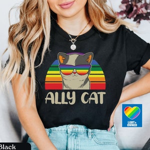Może przedstawiać: Czarny T-shirt z tęczowym zachodem słońca i kotem w tęczowych okularach przeciwsłonecznych. Na koszulce nadrukowany jest napis "Ally Cat". W prawym dolnym rogu zdjęcia widoczna jest naklejka z napisem "LGBT+ Owned".