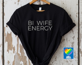 May include: Black t-shirt with white text that reads "Bi Wife Energy". A rainbow heart sticker with the text "LGBT+ Owned" is in the bottom right corner of the image.