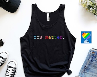 May include: Black tank top with the text "You matter." in rainbow colors. The tank top is laid flat on a white surface with a pair of black sneakers, a black baseball cap, sunglasses, a pair of blue denim shorts, and a sticker with a rainbow heart and the text "LGBT+ Owned".