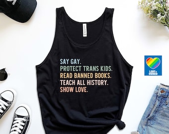 Puede incluir: Camiseta de tirantes negra con el texto "SAY GAY. PROTECT TRANS KIDS. READ BANNED BOOKS. TEACH ALL HISTORY. SHOW LOVE." en colores arcoíris. Un corazón arcoíris con el texto "LGBT+ OWNED" está en la esquina inferior derecha.