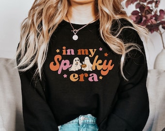 Może przedstawiać: Czarna bluza z napisem "in my Spooky era" w pomarańczowych, różowych i żółtych literach. Projekt zawiera dwa duchy i dynię. Bluza jest artykułem o tematyce Halloween.