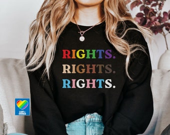Può includere: Una felpa nera con la parola "RIGHTS." ripetuta tre volte in colori arcobaleno. La felpa è indossata da una persona con lunghi capelli biondi. Il testo "LGBT+ OWNED" è visibile su un piccolo adesivo a forma di cuore arcobaleno.