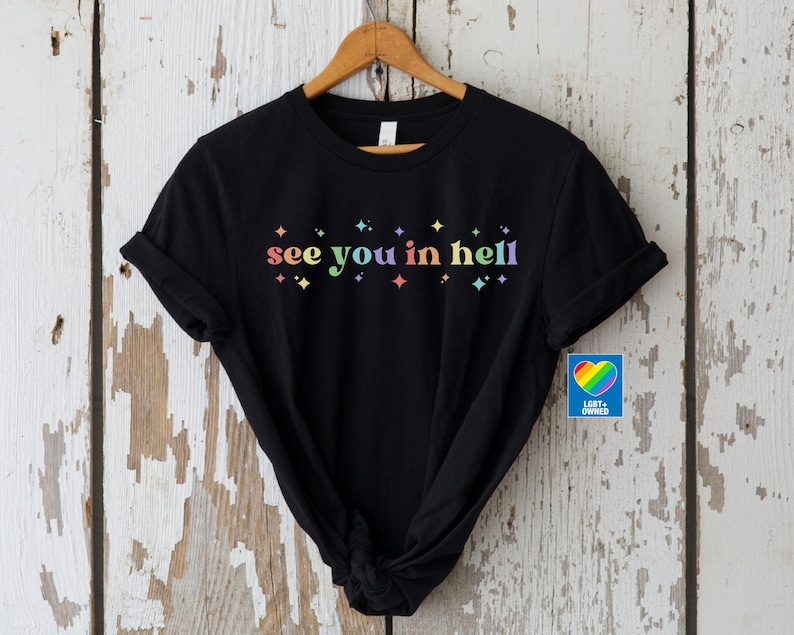 Puede incluir: Camiseta negra con el texto "see you in hell" en colores arco&iacute;ris y peque&ntilde;as estrellas arco&iacute;ris. La camiseta est&aacute; sobre un fondo de madera blanca. Un peque&ntilde;o cuadrado azul con un coraz&oacute;n arco&iacute;ris y el texto "LGBT+ OWNED" est&aacute; en la esquina inferior derecha.