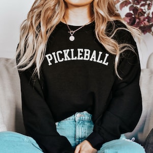 May include: A black sweatshirt with the word "Pickleball" printed in white on the front. The sweatshirt is worn by a person with long blonde hair.