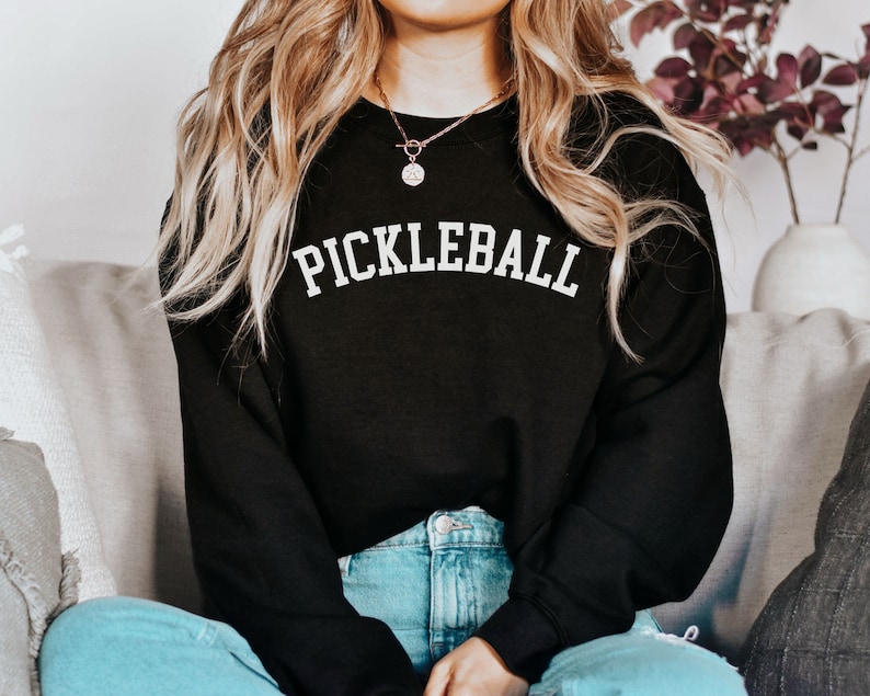 May include: A black sweatshirt with the word "Pickleball" printed in white on the front. The sweatshirt is worn by a person with long blonde hair.