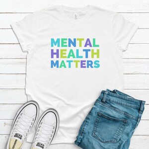 Puede incluir: Camiseta blanca con el texto "MENTAL HEALTH MATTERS" en colores arco&iacute;ris. La camiseta est&aacute; colocada sobre una superficie de madera blanca con un par de zapatillas blancas y unos vaqueros azules.