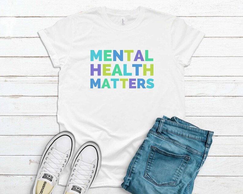 Puede incluir: Camiseta blanca con el texto "MENTAL HEALTH MATTERS" en colores arco&iacute;ris. La camiseta est&aacute; colocada sobre una superficie de madera blanca con un par de zapatillas blancas y unos vaqueros azules.