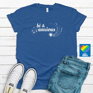 Pode incluir: Uma camiseta azul com as palavras "bi & anxious" em branco, acompanhada por um design de estrela. A camiseta &eacute; combinada com jeans azuis e t&ecirc;nis brancos. Um logotipo de cora&ccedil;&atilde;o arco-&iacute;ris com "LGBT+ OWNED" &eacute; vis&iacute;vel.