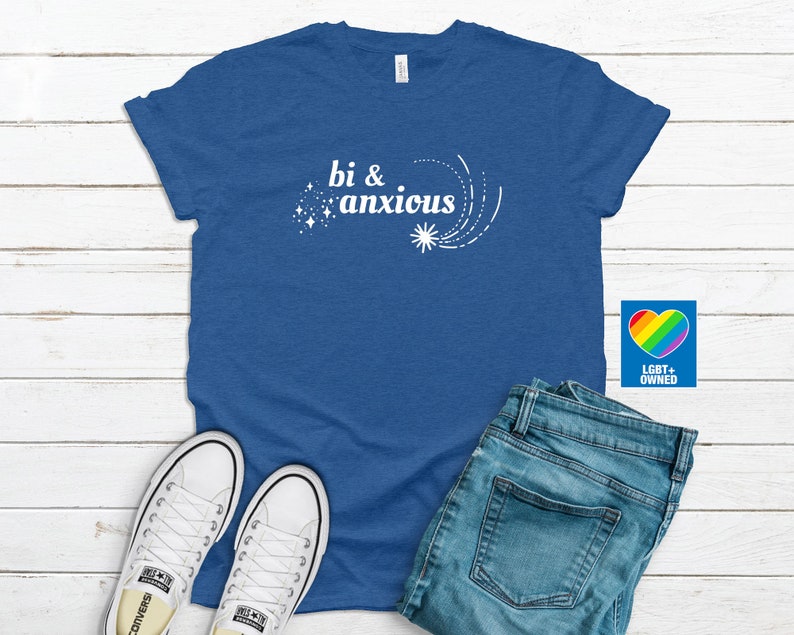 Pode incluir: Uma camiseta azul com as palavras "bi & anxious" em branco, acompanhada por um design de estrela. A camiseta &eacute; combinada com jeans azuis e t&ecirc;nis brancos. Um logotipo de cora&ccedil;&atilde;o arco-&iacute;ris com "LGBT+ OWNED" &eacute; vis&iacute;vel.