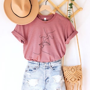 May include: A heather mauve t-shirt with a line drawing of two people kissing. The shirt is on a hanger with a straw hat and a woven bag.
