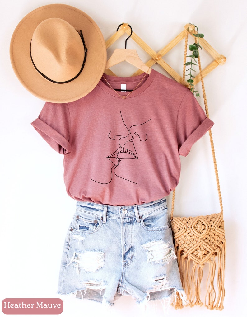 May include: A heather mauve t-shirt with a line drawing of two people kissing. The shirt is on a hanger with a straw hat and a woven bag.