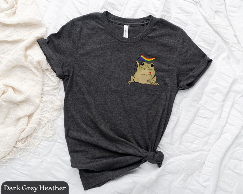 May include: Dark grey heather t-shirt with a cartoon frog wearing a rainbow pride flag. The frog is holding a small red heart.