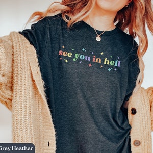 Puede incluir: Camiseta de color gris oscuro jaspeado con el texto "see you in hell" en colores arco&iacute;ris y peque&ntilde;as estrellas blancas.