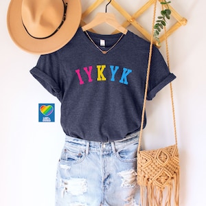 May include: A heather navy blue t-shirt with the text "IYKYK" in rainbow colors. The shirt is hanging on a wooden hanger with a straw hat and a woven bag.