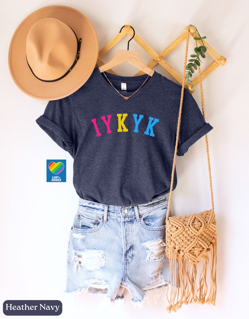 May include: A heather navy blue t-shirt with the text "IYKYK" in rainbow colors. The shirt is hanging on a wooden hanger with a straw hat and a woven bag.