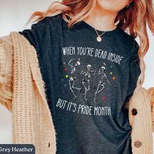 May include: Dark grey heather t-shirt with the text "When you're dead inside but it's pride month" and a graphic of three skeletons dancing. The skeletons are wearing rainbow colors.