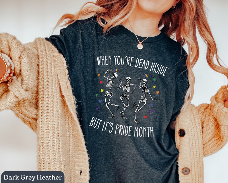 May include: Dark grey heather t-shirt with the text "When you're dead inside but it's pride month" and a graphic of three skeletons dancing. The skeletons are wearing rainbow colors.