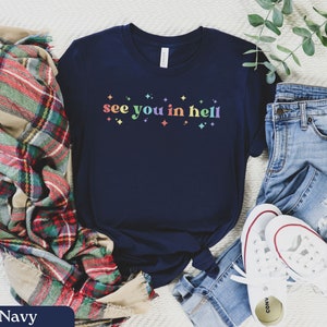Puede incluir: Una camiseta azul marino con el texto "see you in hell" impreso en colores arco&iacute;ris. La camiseta est&aacute; combinada con una bufanda de cuadros, unos vaqueros azules y unas zapatillas blancas.