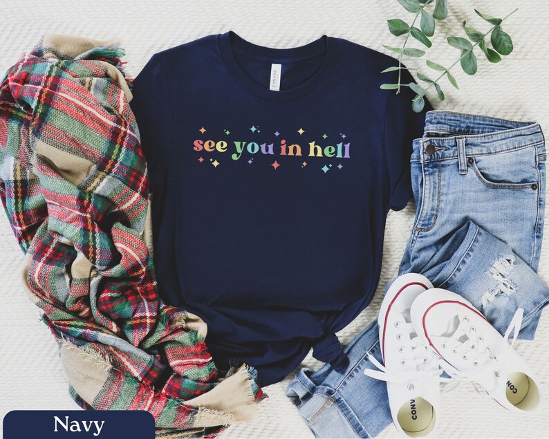 Puede incluir: Una camiseta azul marino con el texto "see you in hell" impreso en colores arco&iacute;ris. La camiseta est&aacute; combinada con una bufanda de cuadros, unos vaqueros azules y unas zapatillas blancas.