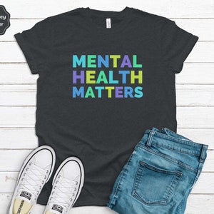 Puede incluir: Camiseta de color gris oscuro jaspeado con el texto "MENTAL HEALTH MATTERS" en colores arco&iacute;ris.