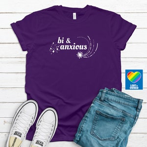 Pode incluir: Uma camiseta roxa com o texto branco "bi & anxious" e um design de estrela. A imagem inclui jeans azuis e t&ecirc;nis brancos. Um logotipo de cora&ccedil;&atilde;o arco-&iacute;ris com "LGBT+ OWNED" tamb&eacute;m &eacute; vis&iacute;vel.
