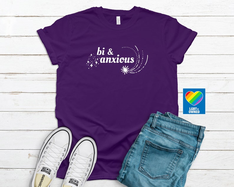 Pode incluir: Uma camiseta roxa com o texto branco "bi & anxious" e um design de estrela. A imagem inclui jeans azuis e t&ecirc;nis brancos. Um logotipo de cora&ccedil;&atilde;o arco-&iacute;ris com "LGBT+ OWNED" tamb&eacute;m &eacute; vis&iacute;vel.