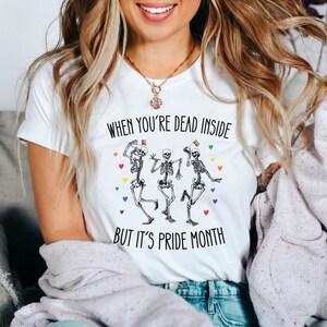 May include: White t-shirt with the text "When you're dead inside but it's pride month" and a graphic of three skeletons dancing with rainbow flags.