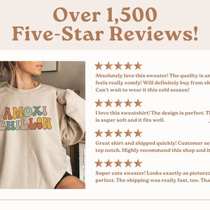 May include: A beige sweatshirt with the text "AMOXI CHILLON" in colorful retro font. The sweatshirt has a crew neck and long sleeves. The model is wearing the sweatshirt and smiling.