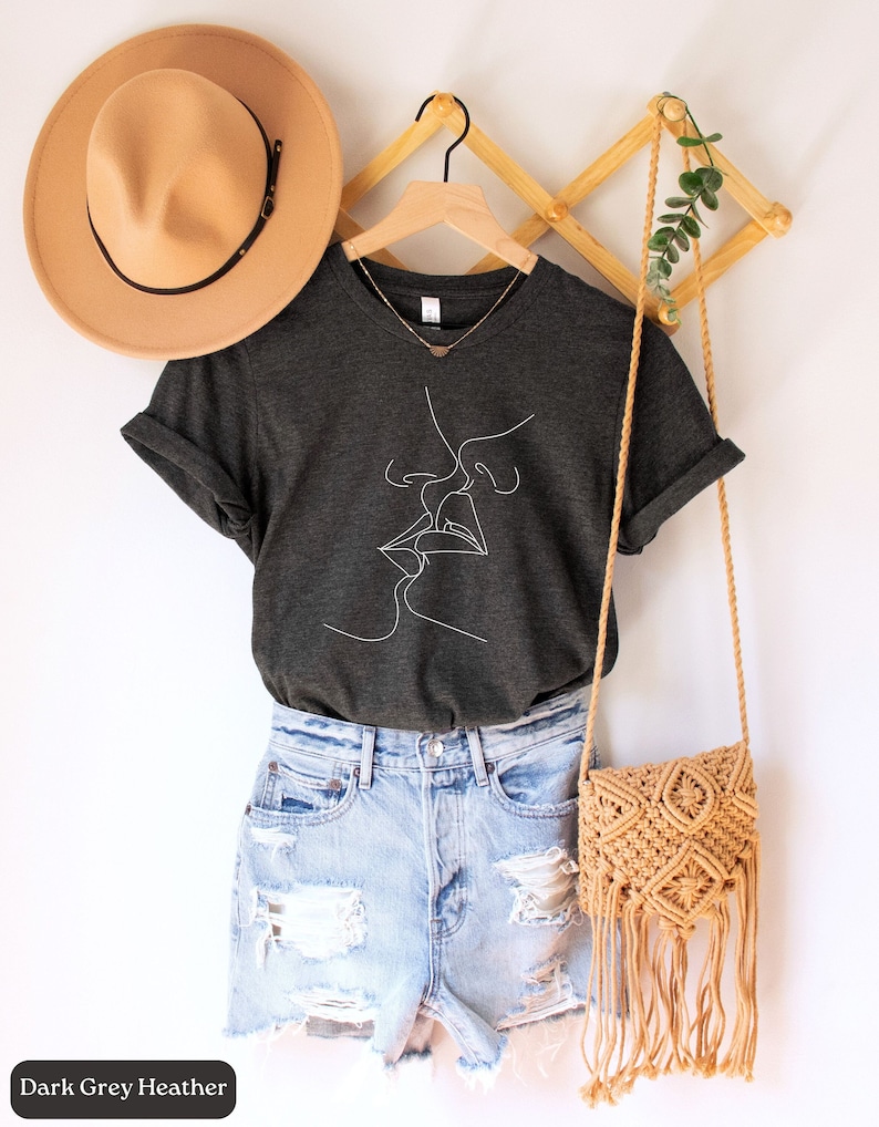 May include: A dark grey heather t-shirt with a white line drawing of two people kissing. The shirt is hanging on a wooden hanger with a brown hat and a woven bag.