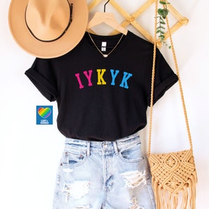 May include: Black t-shirt with the text "IYKYK" in rainbow colors. The shirt is on a hanger and is being modeled by a person wearing denim shorts and a straw hat.