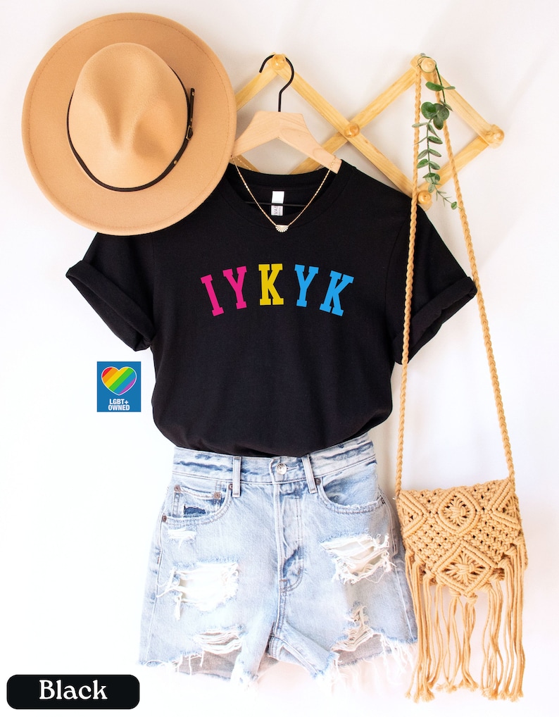 May include: Black t-shirt with the text "IYKYK" in rainbow colors. The shirt is on a hanger and is being modeled by a person wearing denim shorts and a straw hat.