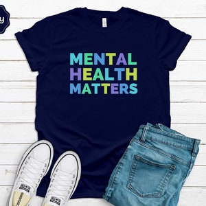 Puede incluir: Una camiseta azul marino con el texto "MENTAL HEALTH MATTERS" en colores arco&iacute;ris.