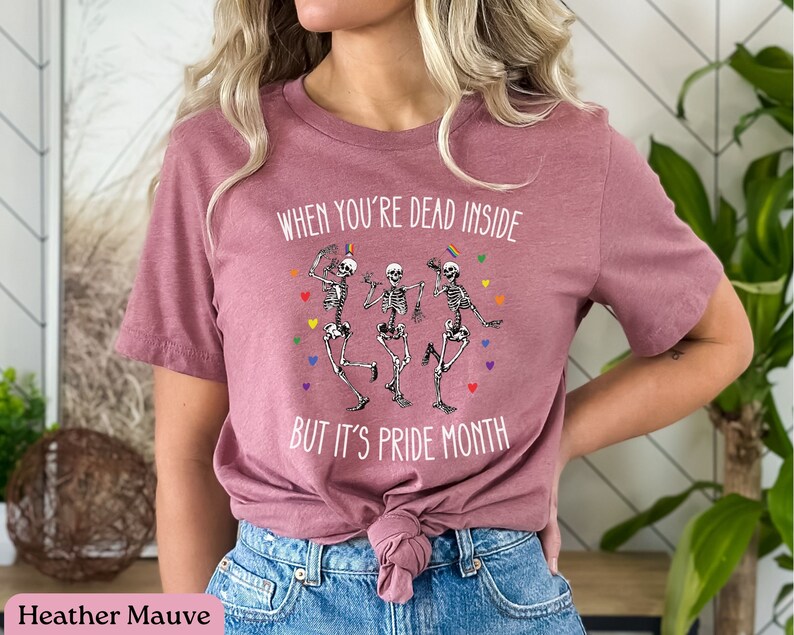 May include: Heather mauve t-shirt with a graphic of three skeletons dancing with rainbow colors and the text "When you're dead inside but it's pride month."