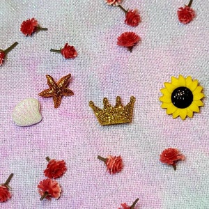 May include: A collection of small, decorative items including a gold crown, a white heart, a yellow sunflower, and a gold starfish. The items are arranged on a pink and white fabric background.