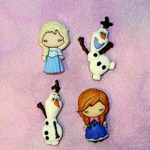 Puede incluir: Cuatro botones de personajes con personajes de la película Frozen. Los botones son blancos, azules, marrones y azul claro. Los personajes son Elsa, Olaf, Anna y Olaf.