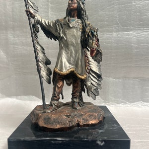 May include: A silver and bronze sculpture of a Native American man wearing a headdress with feathers and holding a spear. The sculpture is on a black marble base and is signed "The Covenant" by C.A. Pade.