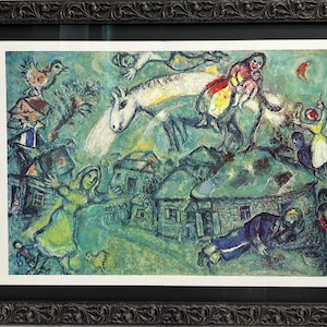 Pode incluir: Uma impressão emoldurada de uma pintura colorida e fantasiosa. A obra de arte retrata uma cena de vila com figuras, animais e um cavalo, todos renderizados em um estilo onírico. A paleta de cores inclui azuis, verdes e vermelhos, com uma moldura preta ornamentada.
