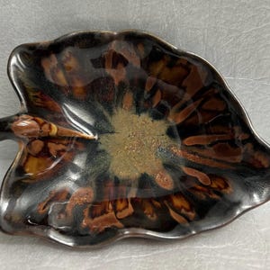 May include: A brown and black ceramic leaf-shaped dish with a textured glaze. The dish has a raised edge and a small handle on one side.