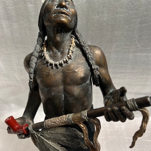 May include: Bronze sculpture of a Native American man holding a pipe, with feathers and beads. The sculpture is on a black base.