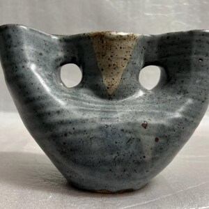 Può includere: Un vaso in ceramica grigia con una forma astratta unica. Ha due fori su entrambi i lati e una glassa bianca e marrone sulla parte superiore.