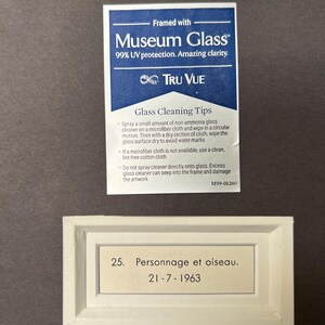 May include: A white framed label with black text that reads "25. Personnage et oiseau. 21-7-1963". The label is on a dark brown background.
