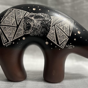 Pode incluir: Escultura de urso em cerâmica com corpo preto e pernas castanhas. A cabeça do urso apresenta uma gravura detalhada de um urso a rugir e padrões geométricos. A escultura tem o texto "Dinch" e "Harrison Juan".
