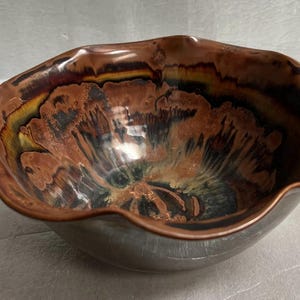 May include: A brown ceramic bowl with a wavy rim and a colorful, abstract design. The design features shades of brown, black, yellow, and white.