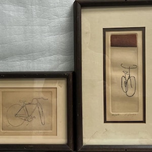 May include: Two framed black and white line drawings of bicycles. The drawing on the left shows a bicycle with two wheels and a handlebar. The drawing on the right shows a bicycle with two wheels and a handlebar. Both drawings are in simple lines and are framed in black wood frames.