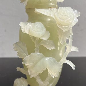 Op de afbeelding: Een lichtgroene, gebeeldhouwde stenen vaas met een bloemenontwerp. De vaas heeft gedetailleerde gravures van rozen en bladeren, waarbij de bloemen en het gebladerte zich uitstrekken vanaf het oppervlak van de vaas. De algehele esthetiek is delicaat en sierlijk.