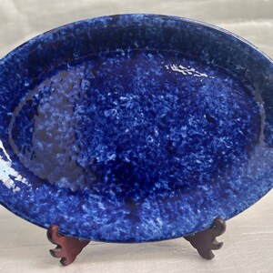 Könnte beinhalten: Eine blaue ovale Keramik-Servierplatte mit einem marmorierten Überzug. Die Platte steht auf einem Holzständer.