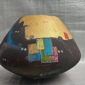 May include: A hand-painted, abstract ceramic vase with a dark brown base and a gold-leaf top. The vase features colorful geometric shapes in blue, green, pink, and yellow. Red stripes and white diamond shapes add detail to the design.