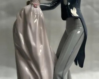 May include: A porcelain figurine of a couple dancing. The woman is wearing a long, flowing pink dress and the man is wearing a black suit with a white shirt and a black bow tie. The figurine is in a romantic pose, with the man holding the woman's hand and the woman looking up at him.