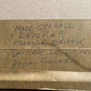 May include: A close-up of a brown paper label with handwritten text. The text reads "Marc Chagall, R&ecirc;verie, Maeght Editeur, 1969, Derriere Le Miroir, Paris France."